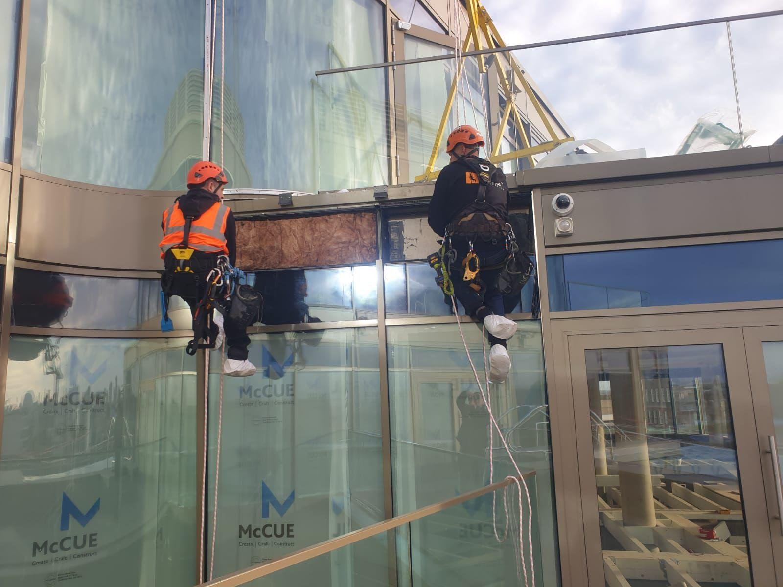 Two rope-access technicians replacing glazing on a curved glass façade