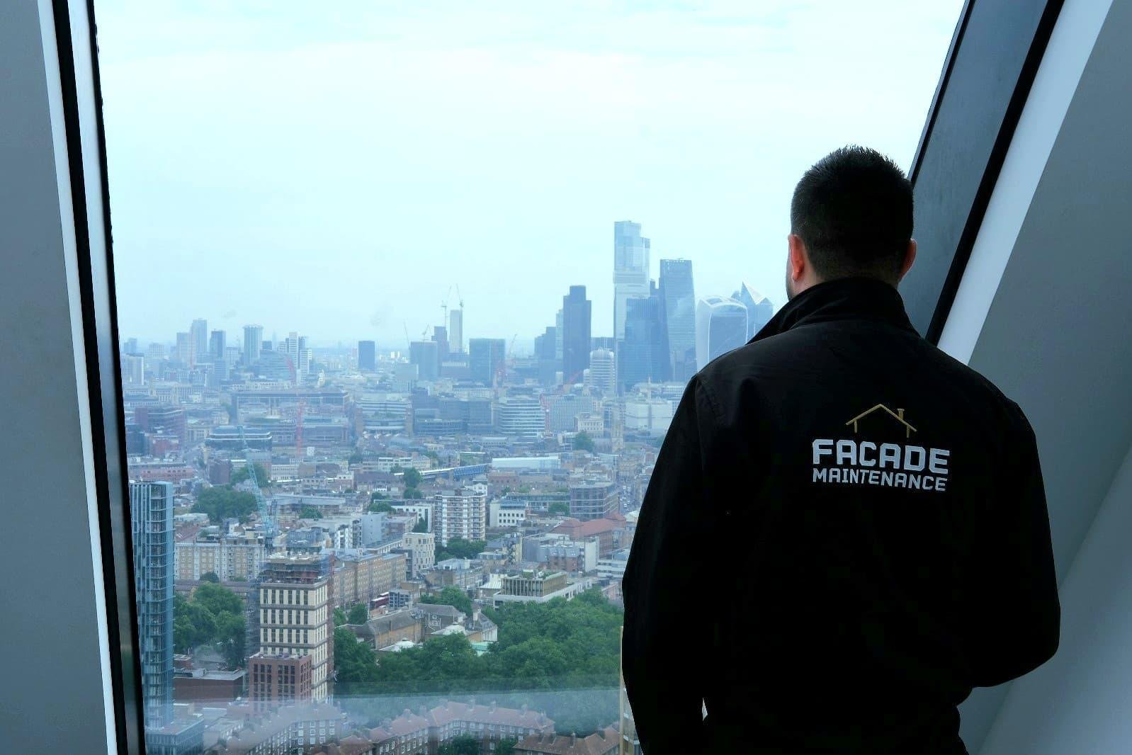Facade Maintenance team member in branded jacket looking out over the City of London skyline from a high-rise